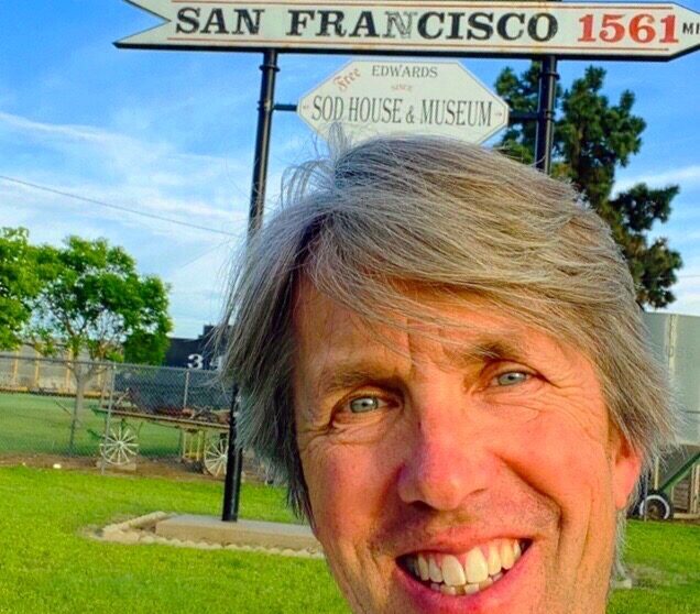 A person takes a selfie in front of a sign that says, "New York: 1,561 miles and San Francisco 1,561 miles." They are at the Midway Point of the U.S.A in Kinsley, Kansas.